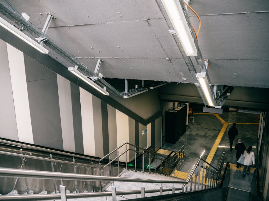 Inside a multi-level underground station with a view of a staircase leading downwards. The staircase has metallic handrails and steps with anti-slip yellow strips along the edges. Overhead, there are